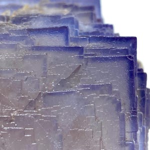 May include: Close-up of a layered, crystalline structure. The mineral formation features a gradient of colors, transitioning from a pale, translucent base to deep blue edges. The geometric shapes create a textured, almost architectural appearance.