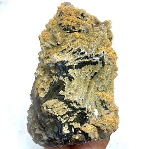 May include: A natural mineral specimen featuring a layered, dark gray and black base with a textured, light tan and white coating. The rough-hewn surface displays a crystalline structure, suitable for display or collection.