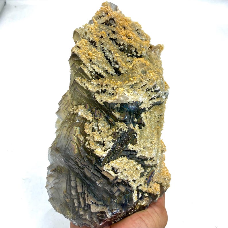 May include: A large, raw fluorite crystal with a complex, layered structure. The crystal features a mix of gray, brown, and clear sections, with a coating of light tan, textured material on top. The overall shape is irregular and natural.
