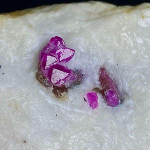 May include: A raw mineral specimen featuring vibrant magenta-colored crystal clusters embedded within a textured, opaque white matrix. The crystalline formations exhibit sharp, angular facets and a deep, saturated pink hue contrasted against the pale stone.
