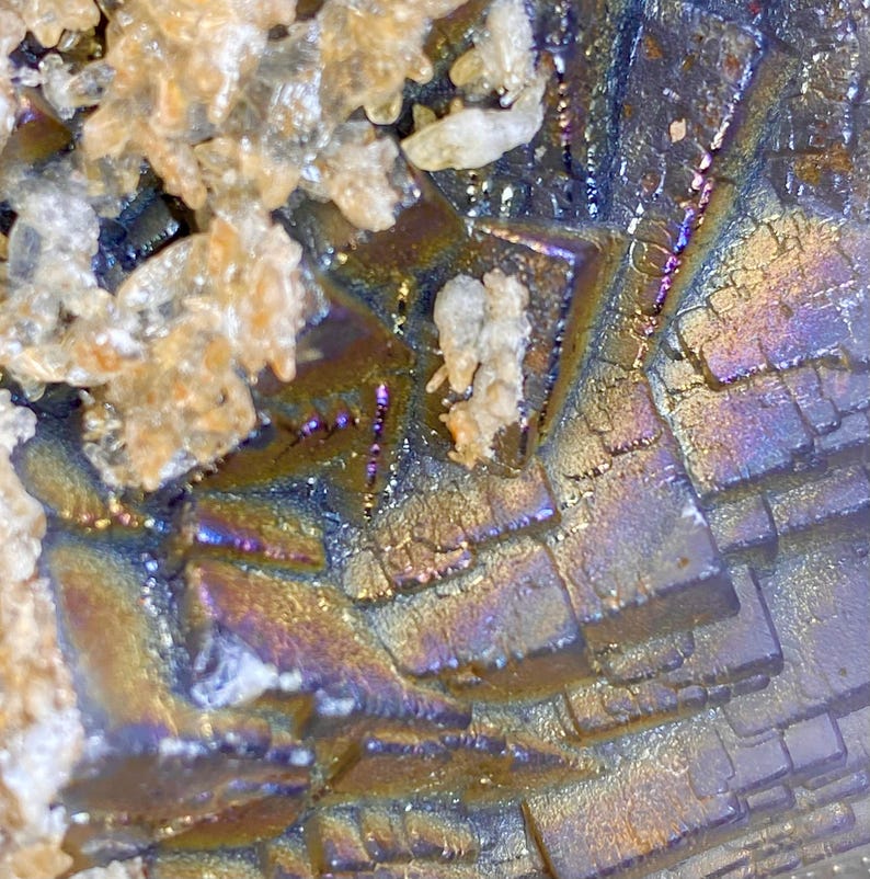 May include: Close-up of a mineral formation with a metallic sheen. The surface displays a mosaic of geometric shapes in shades of blue, purple, gold, and brown. Some areas have a crystalline texture, with small, light-colored formations.