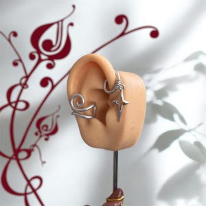 May include: Silver ear cuffs and a star-shaped earring are displayed on a mannequin ear. The ear cuffs have a spiral and a curved design. The star earring hangs from a hoop. The background is white with red floral accents.