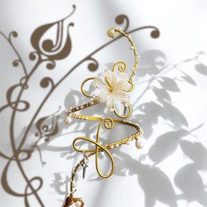 May include: A gold-toned arm cuff with a floral design. The cuff features a white flower with pearl accents, and a dangling cross charm. The design is intricate and elegant, with a delicate, feminine aesthetic.