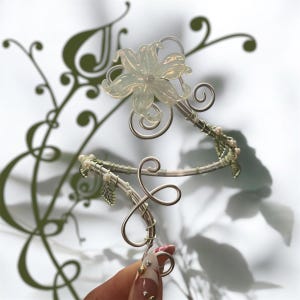 May include: A delicate silver-toned floral hair accessory featuring a translucent flower and intricate wirework. The design includes spiraling accents and small leaf details, creating an elegant, nature-inspired look. The accessory is held by a hand with a manicure.