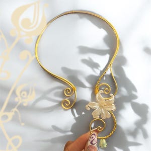 Puede incluir: Un collar dorado con un acento floral. El collar presenta un diseño delicado y curvo con una flor y pequeños acentos verdes. El collar es sostenido por una mano con un diseño de uñas pintadas.