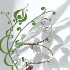 May include: A silver metal wine bottle holder in a decorative S-shape. It has small green leaf accents and a swirling design. The background features a green floral pattern and shadows.