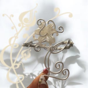 Peut inclure: Deux pinces à cheveux argentées. L'une présente un motif floral avec un centre translucide. L'autre a un design décoratif et tourbillonnant. Les pinces sont tenues par une main avec une manucure, sur fond blanc.