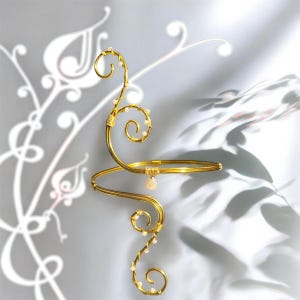 May include: A gold-toned arm cuff with a swirling, wire-wrapped design. The cuff features a spiral element and is adorned with small, pearl-like beads. A small, teardrop-shaped charm hangs from the cuff. The background is a soft, light gray with a subtle floral pattern.