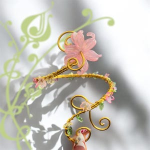 May include: A gold arm cuff with a large pink flower and smaller pink and green floral accents. The cuff features delicate wire detailing and a curved design, creating an elegant, nature-inspired accessory.