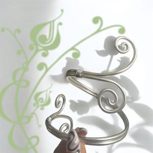 May include: A silver arm cuff with a flowing, open design. The cuff has multiple loops and curves, with a wrapped section at the top. The background is white with a green floral pattern.