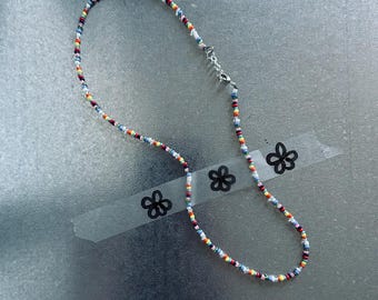 May include: A colorful beaded necklace with a silver clasp. The necklace features small, round beads in various colors, including red, yellow, blue, and white. The necklace is displayed on a gray surface with flower-shaped stickers.