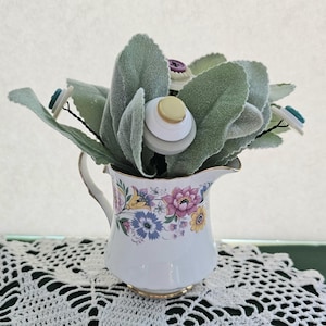 May include: A decorative floral arrangement in a vintage white pitcher. The pitcher is adorned with a floral pattern in pink, yellow, and blue. The arrangement features large, soft green leaves and button accents in various colors.