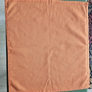 May include: A square, orange-colored fabric napkin with stitched edges. The napkin is made of a plain weave fabric and is displayed on a green surface. The napkin is a solid color and has a simple, classic design.