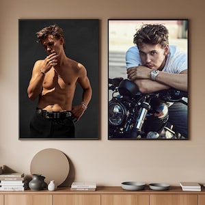 Può includere: Due stampe artistiche incorniciate. Una presenta una persona a torso nudo con pantaloni neri, l'altra una persona con una maglietta bianca appoggiata a una motocicletta. Entrambe le stampe hanno cornici nere e sono esposte su una parete beige.