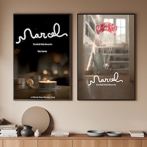 Puede incluir: Dos carteles de películas enmarcados. El cartel de la izquierda tiene un fondo negro con el título "Marcel" en escritura blanca, y el texto "The Shell With Shoes On" y "The Movie". El cartel de la derecha muestra un personaje con un paracaídas.