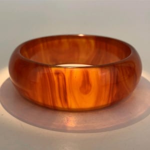 May include: A wide, round bangle bracelet in shades of amber and orange. The translucent material has a marbled appearance with vertical streaks. The bracelet is sitting on a light pink surface.