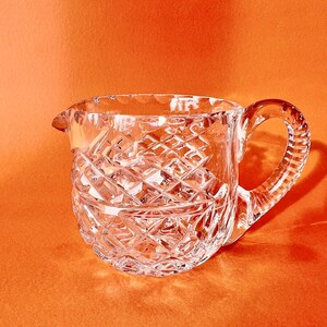 May include: A clear, cut-glass creamer with a diamond pattern and a handle. The creamer has a spout for easy pouring. The handle has a textured design. The creamer is on an orange background.