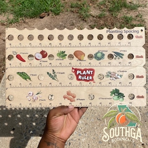 May include: A wooden plant ruler with planting spacing measurements in inches. The ruler features illustrations of various vegetables and the text "Plant Ruler". The ruler is held by a person.