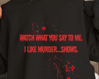 Puede incluir: Sudadera negra con el texto rojo "WATCH WHAT YOU SAY TO ME. I LIKE MURDER...SHOWS." y detalles de salpicaduras rojas. Una prenda de manga larga de estilo casual.