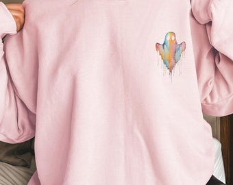 Puede incluir: Una sudadera rosa claro con un diseño de fantasma en acuarela en la parte superior izquierda del pecho. El fantasma es una mezcla de colores arcoíris con efectos de pintura goteando. La sudadera tiene mangas largas y cuello redondo.