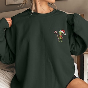 Könnte beinhalten: Dunkelgrünes Sweatshirt mit einem gestickten Alien, das einen Weihnachtsmannhut trägt und eine Zuckerstange hält. Das Alien ist grün mit schwarzen Augen und einem roten Schal. Das Sweatshirt ist einfarbig und hat einen Rundhalsausschnitt.