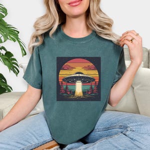 May include: Teal green t-shirt featuring a retro-style graphic of a UFO emitting a beam of light. The design includes a sunset backdrop with trees, clouds, and a starry sky. The shirt is made of a soft material.