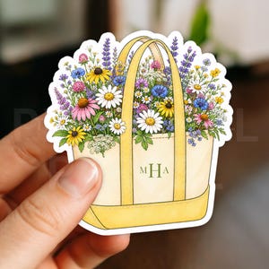 May include: A sticker featuring a cream-colored tote bag with yellow handles and trim, filled with a variety of colorful wildflowers. The bag has the initials "MHA" printed on it, and the sticker is held by a hand.