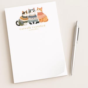May include: A white notepad with a watercolor illustration of three cats wearing sweaters. The cats are in shades of orange, gray, and black. The text "Catwalk Certified - sweater edition" is printed in gold.