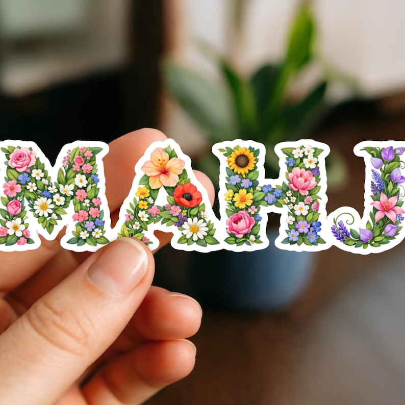 May include: A floral sticker spelling out the word "MAHJ". Each letter is formed with a variety of colorful flowers, including roses, sunflowers, and daisies. The sticker is held between fingers against a blurred background.