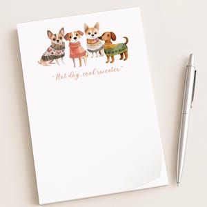 May include: A white notepad with a watercolor illustration of four dogs wearing sweaters. The text "Hot dog, cool sweater" is written in a cursive font. A silver pen rests on the right side of the notepad.