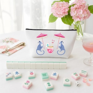 Könnte beinhalten: Eine weiße Kosmetiktasche mit schwarzem Reißverschluss, verziert mit einem skurrilen Design von blauen Affen unter rosa Sonnenschirmen. Mahjong-Steine und ein Satz Steine sind vor der Tasche angeordnet, mit einem rosa Cocktail und Blumen im Hintergrund.