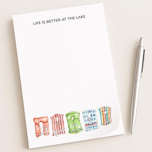 May include: A white notepad with the text "LIFE IS BETTER AT THE LAKE" at the top. The bottom of the notepad features a watercolor illustration of five colorful life vests. A silver pen rests on the right side of the notepad.
