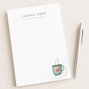 May include: A white notepad with the text "COFFEE FIRST" in black and a teal watercolor design. A teal coffee cup with a red heart is in the bottom right corner. A silver pen rests on the right side of the notepad.