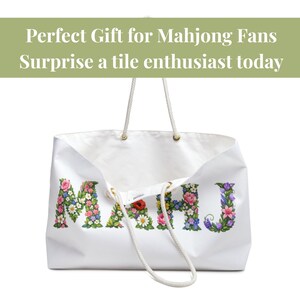 May include: White tote bag with the word "MAHJ" spelled out in colorful floral letters. The bag has rope handles and gold-colored eyelets. Text at the top reads "Perfect Gift for Mahjong Fans Surprise a tile enthusiast today."