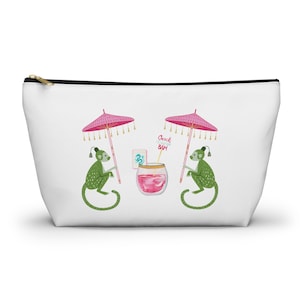 May include: White zippered pouch featuring a whimsical design. Two green monkeys with pink parasols flank a pink cocktail with the text "Beach Bum". The pouch is ideal for cosmetics or travel.