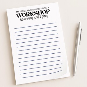 May include: White notepad with blue lined pages and the text "MY HUSBAND AND I ARE DOING A WORKSHOP he works and I shop" in black. A silver pen rests on the right side of the notepad.