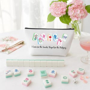 May include: A white zippered pouch with a black zipper, decorated with colorful Mahjong tile illustrations and the text "I Came for the Snacks Stayed for the Mahjong." Several Mahjong tiles and a set of tiles are arranged in front.