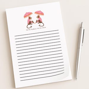 May include: A white notepad with black horizontal lines and a decorative illustration of two monkeys under pink umbrellas. The text "Not My Monkeys, Not My Circus" is below the illustration. A silver pen rests on the right.