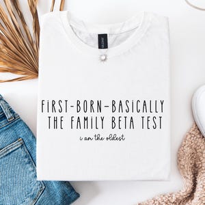 以下が含まれることがあります： 「FIRST-BORN-BASICALLY THE FAMILY BETA TEST」と「i am the oldest」の黒い文字がプリントされた白いTシャツ。Tシャツは折りたたまれ、白い表面に置かれ、青いジーンズとベージュのセーターが見えます。