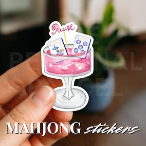 以下が含まれることがあります： 2 つの麻雀牌と「Pause」バナーが付いたピンクのカクテルグラスのステッカー。ステッカーの下部には「MAHJONG stickers」の文字があります。