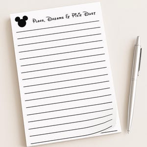 May include: A white notepad with black horizontal lines and the text "Plans, Dreams & Pixie Dust" in black. A black Mickey Mouse head is in the upper left corner. A silver pen rests on the right side of the notepad.