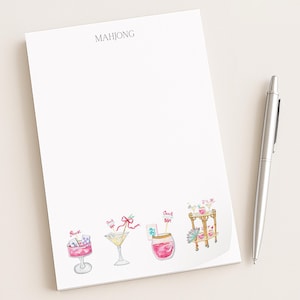 May include: White notepad with the word "MAHJONG" at the top. The notepad features watercolor illustrations of cocktails and a serving cart. A silver pen rests on the right side of the notepad.