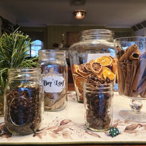 May include: A collection of glass jars filled with various spices and dried ingredients. Includes cloves, bay leaves, dried orange slices, star anise, and cinnamon sticks. The jars are arranged on a patterned tablecloth, creating a rustic and inviting display.