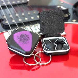 May include: A black and purple guitar pick holder with a purple Dunlop pick that reads "USA 1.14 mm". The holder is open, revealing black earplugs. The items are on a red electric guitar.