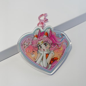 Könnte beinhalten: Ein herzförmiger Acryl-Schlüsselanhänger mit einer Zeichentrickfigur mit rosa Haaren und einer Katze. Die Figur trägt ein weißes und orangefarbenes Outfit. Der Schlüsselanhänger hat einen rosafarbenen Verschluss und einen hellblauen Rand.