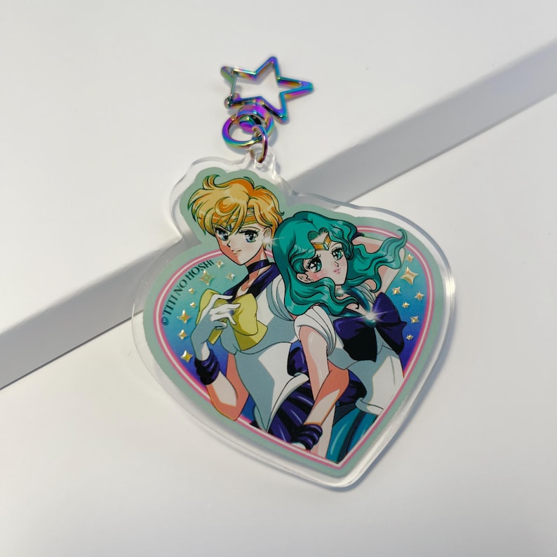 Może przedstawiać: Przezroczysty akrylowy brelok do kluczy z dwoma postaciami z anime w mundurach marynarskich, na tle w kształcie serca. Brelok ma zapięcie w kształcie gwiazdy i srebrny pierścień. Postacie mają blond i turkusowe włosy.