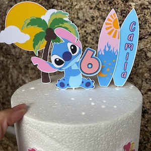 May include: A cake topper featuring a blue cartoon character, a palm tree, a sun, and two surfboards. The surfboards have the name "Camila" written on them. The number "6" is also included, suggesting a birthday theme.