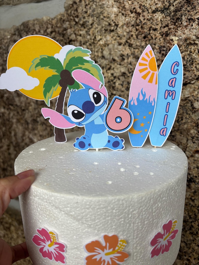 May include: A cake topper featuring a blue cartoon character, a palm tree, a sun, and two surfboards. The surfboards have the name "Camila" written on them. The number "6" is also included, suggesting a birthday theme.