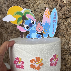 May include: A cake topper featuring a cartoon character, a palm tree, and a sun. The number 6 is displayed, along with two surfboards with the name "Camila". The cake is decorated with colorful hibiscus flowers.
