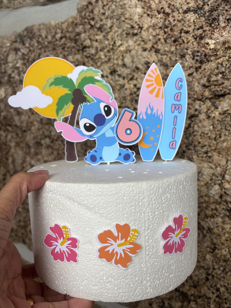 May include: A cake topper featuring a cartoon character, a palm tree, and a sun. The number 6 is displayed, along with two surfboards with the name "Camila". The cake is decorated with colorful hibiscus flowers.